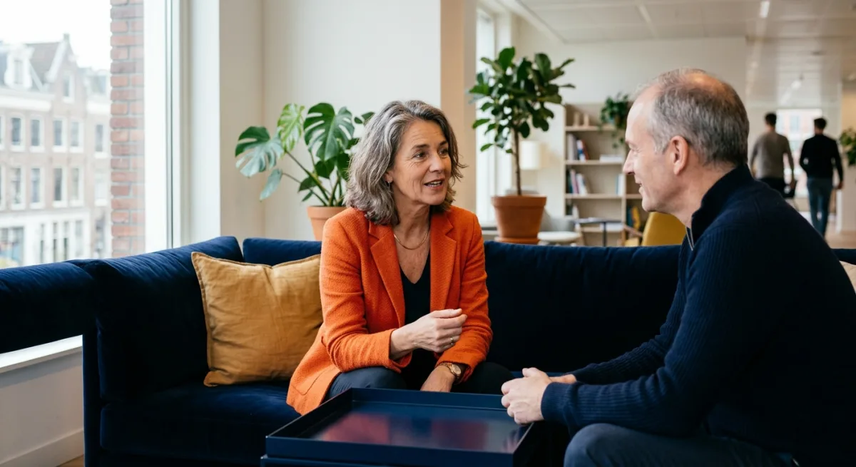 A European executive in an orange blazer talking with a colleague on a navy sofa in an Amsterdam office lounge, illustrating LinkedIn Thought Leader Ads